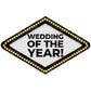 A diamond-shaped marquee sign with the text 'WEDDING OF THE YEAR!' in bold black letters on a white background, surrounded by a black border and glowing warm yellow light bulbs.