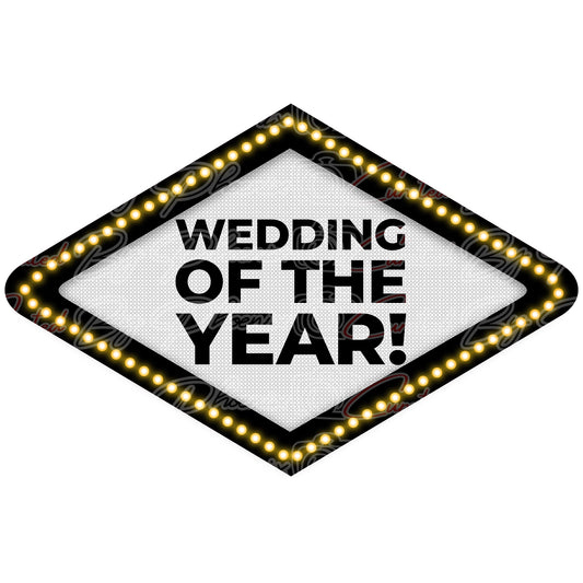 A diamond-shaped marquee sign with the text 'WEDDING OF THE YEAR!' in bold black letters on a white background, surrounded by a black border and glowing warm yellow light bulbs.